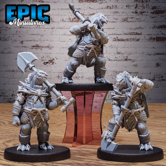 three miniature figures of fantasy creatures, each holding various weapons, standing on a platform against a brick wall background.