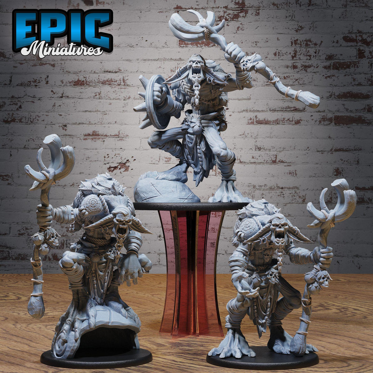 three figurines of fantastical creatures, including a large, imposing figure with a long weapon and a smaller, more detailed figure with a smaller weapon. The figurines are displayed on a wooden surface against a brick wall background.