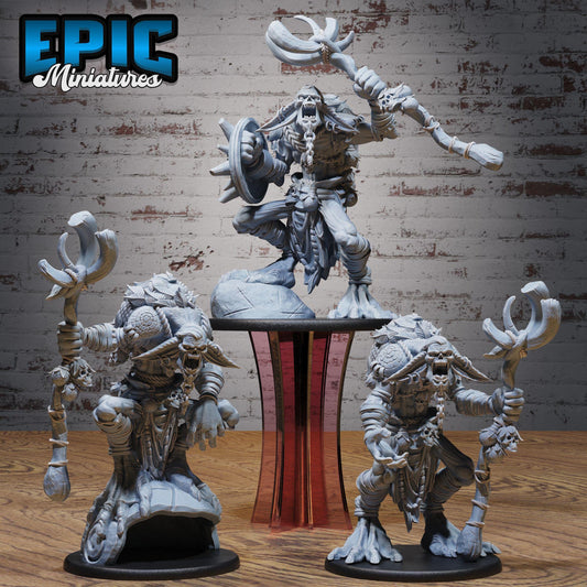 three figurines of fantastical creatures, including a large, imposing figure with a long weapon and a smaller, more detailed figure with a smaller weapon. The figurines are displayed on a wooden surface against a brick wall background.