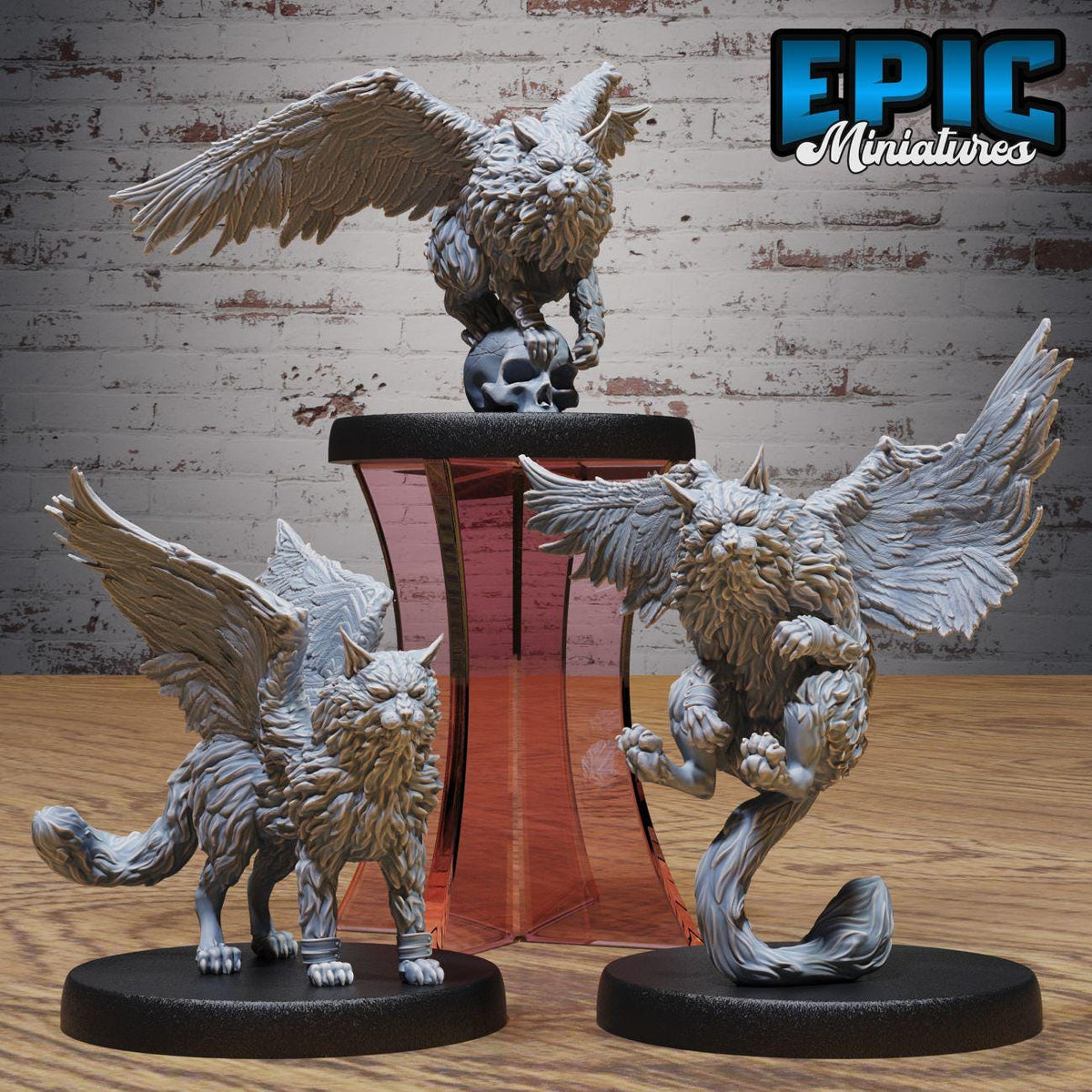 three miniature figurines of winged creatures, including a winged cat and two winged owls, displayed on a stand against a brick wall background.
