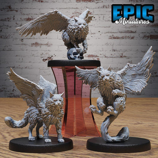 three miniature figurines of winged creatures, including a winged cat and two winged owls, displayed on a stand against a brick wall background.